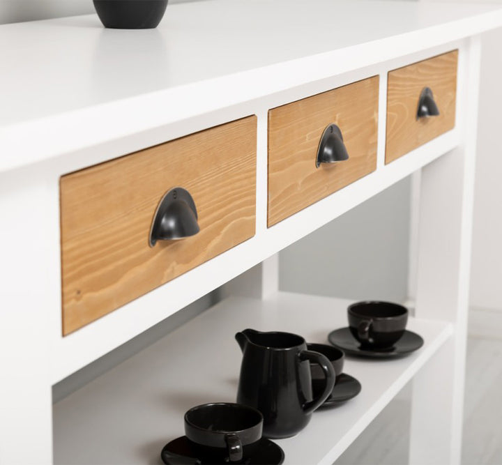 Détail des façades en bois naturel des trois tiroirs avec poignées métalliques sur meuble bas de cuisine en pin massif.