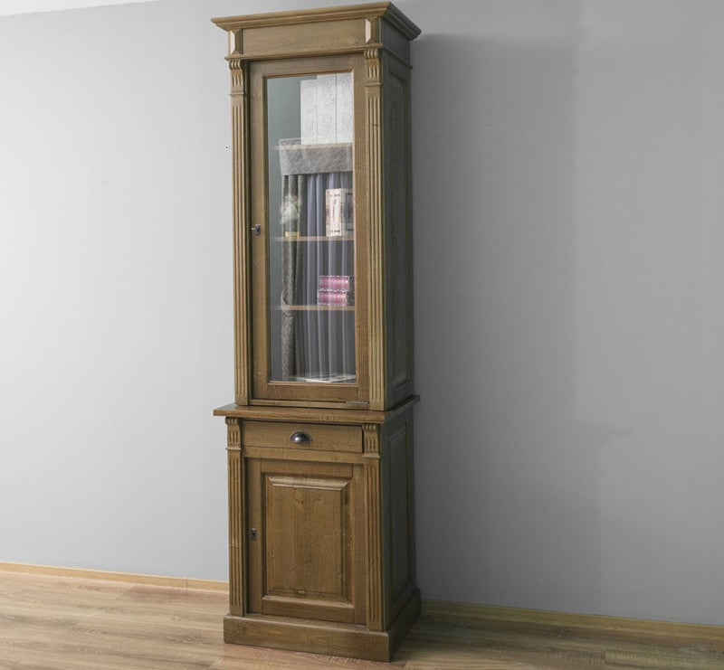 Vitrine en pin massif avec porte vitrée fermée, tiroir central et porte basse, présentée en situation intérieure.